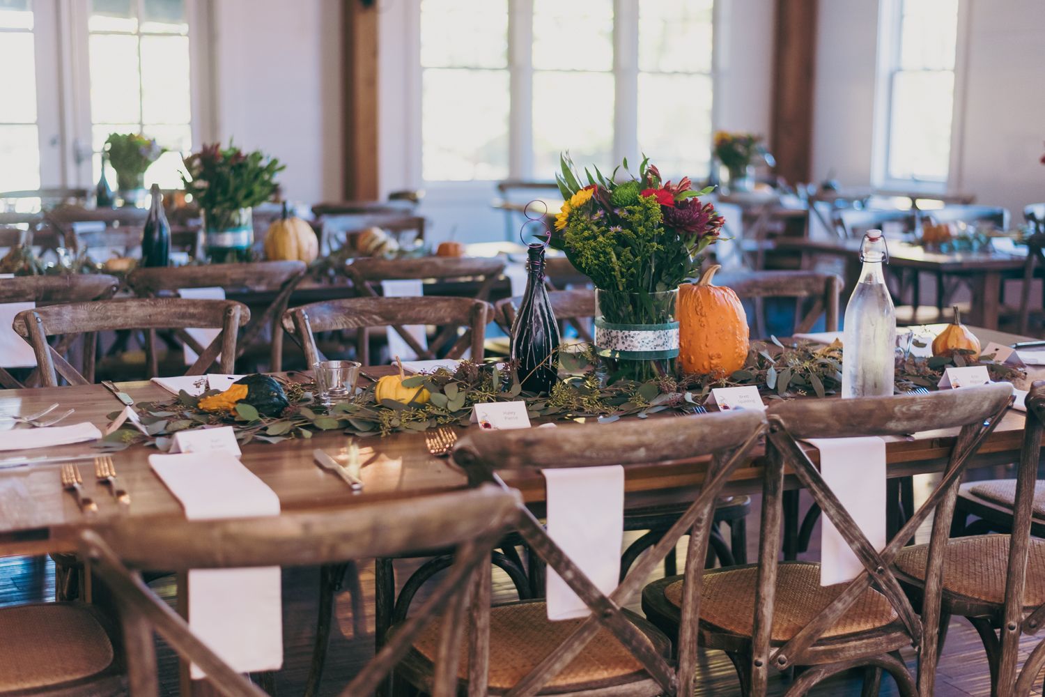Rustic fall wedding reception tables with wooden cross-back chairs, pumpkin and floral centerpieces in a bright venue.