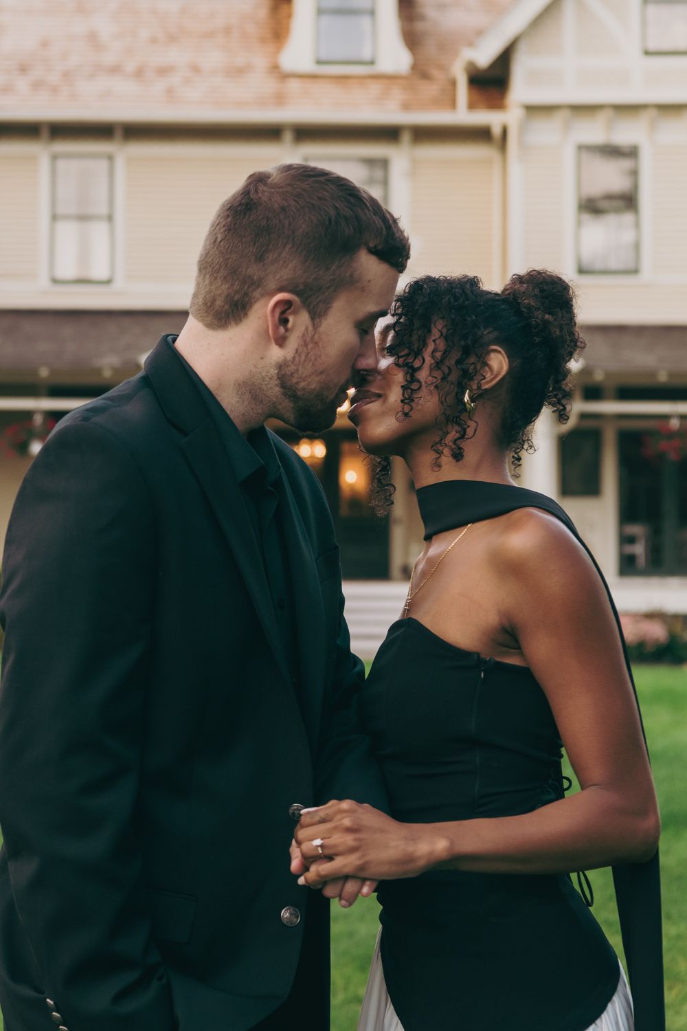 Interracial couple in elegant attire sharing a romantic moment outside a historic house.
