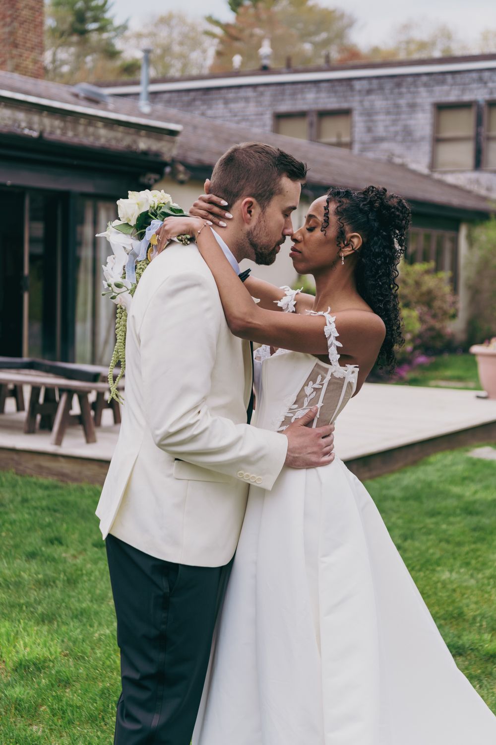 A bride and groom share a romantic kiss outdoors at their wedding venue, surrounded by lush greenery.