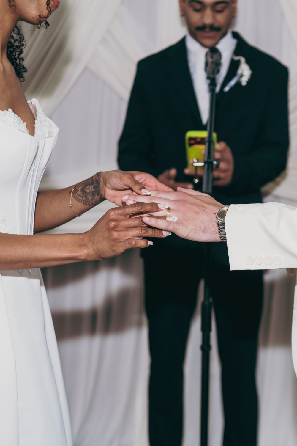 Bride and groom exchange wedding rings during ceremony while officiant holds phone in background.