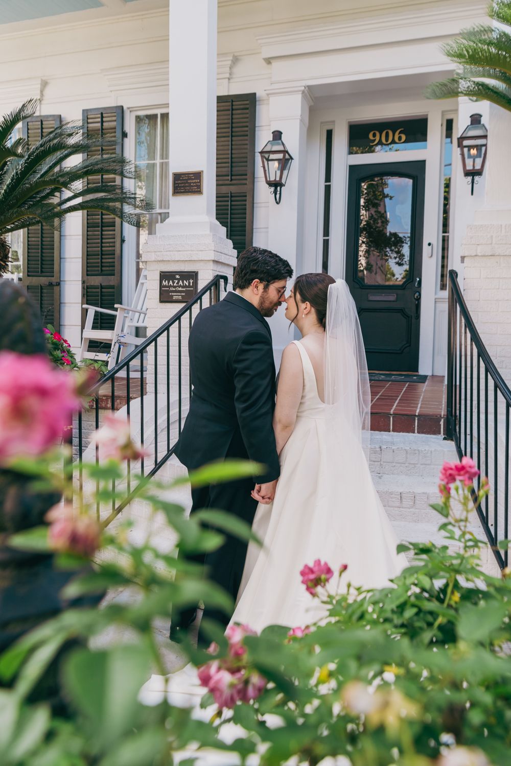 A bride and groom share a romantic moment outside a historic white building at 906, surrounded by blooming flowers.