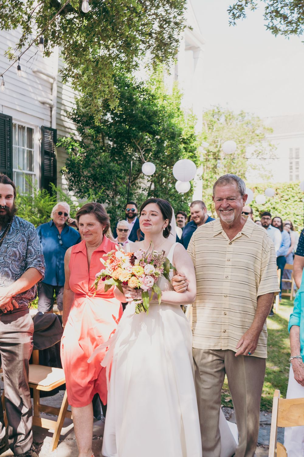 Bride walks down the aisle with parents holding a colorful bouquet at an outdoor garden wedding.