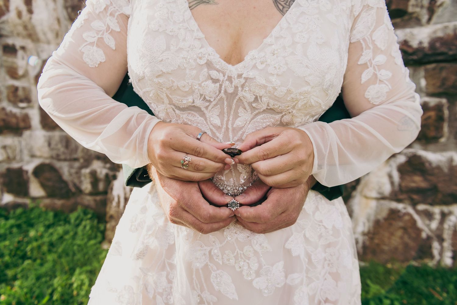 Bride in lace wedding dress holding a decorative brooch, showcasing rings and dark nail polish against a stone wall.