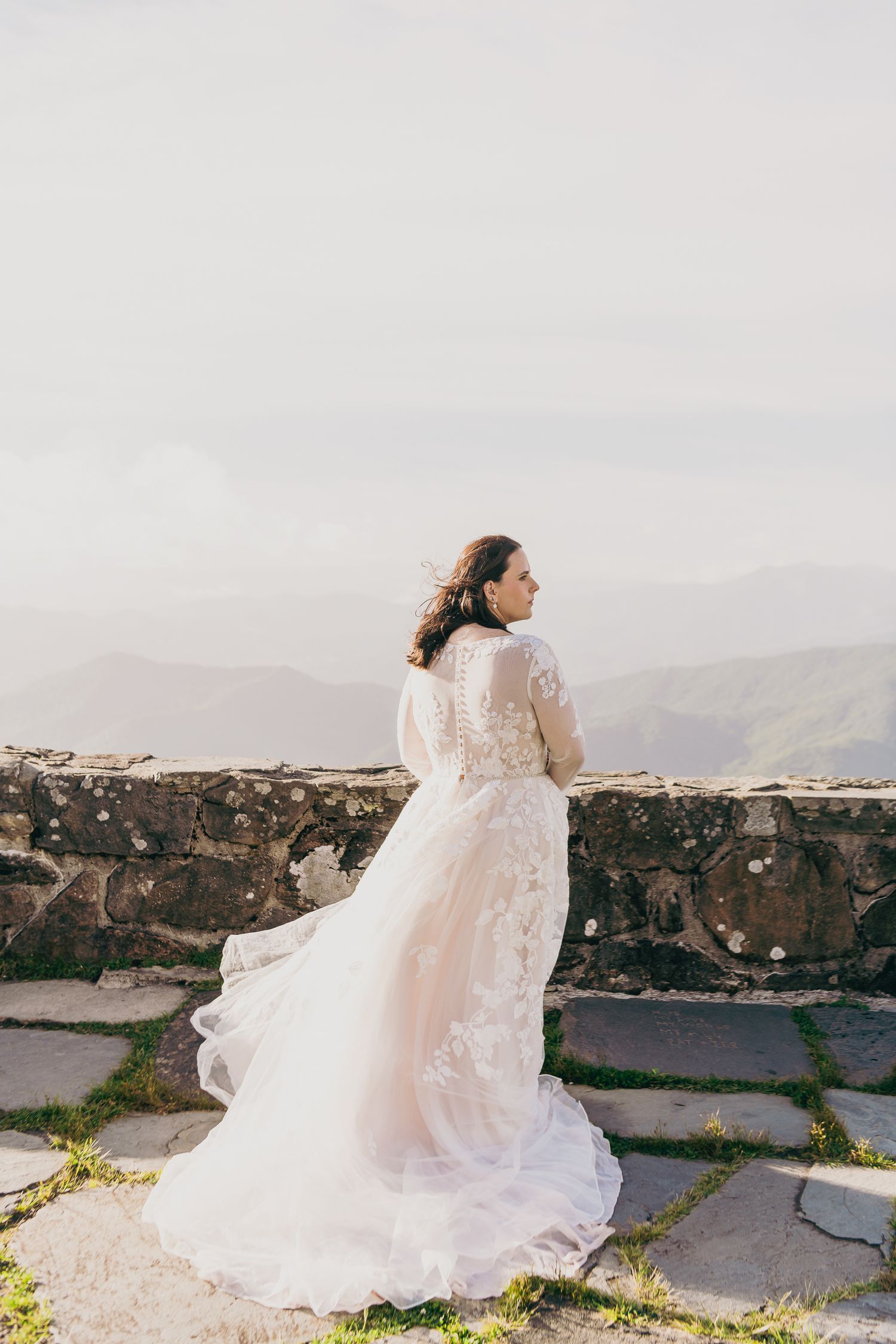 Bride in lace wedding gown with flowing train stands at stone wall overlooking misty mountain landscape.