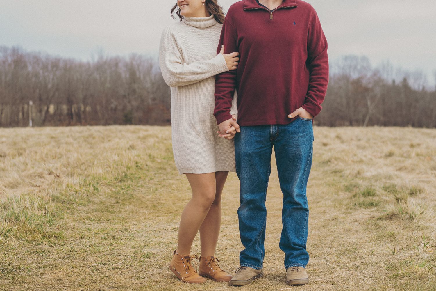 Couple holding hands in a field, woman in white dress and boots, man in red sweater and jeans, bare trees background.