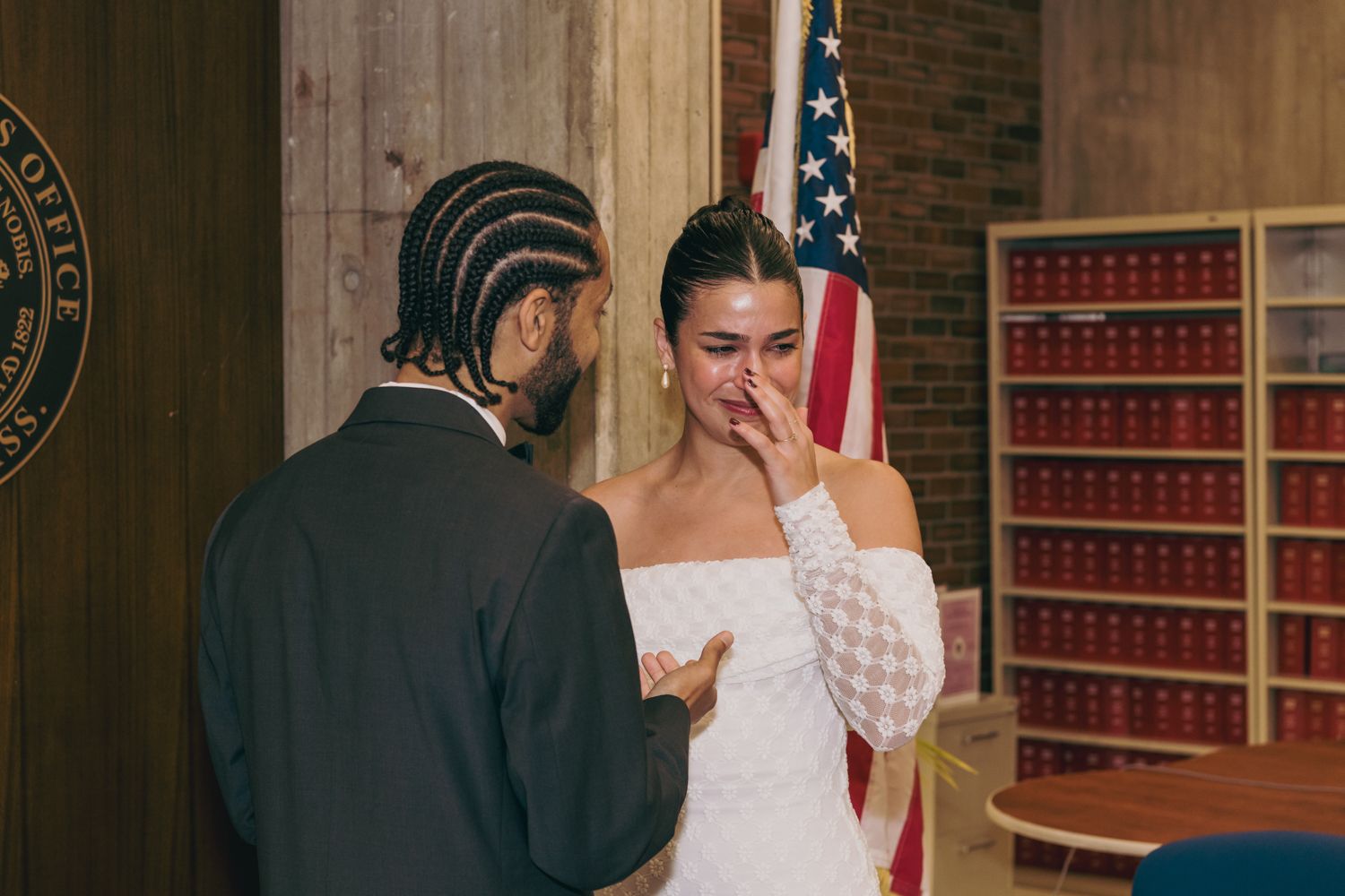 Emotional bride in white dress crying during courthouse wedding ceremony with American flag in background.