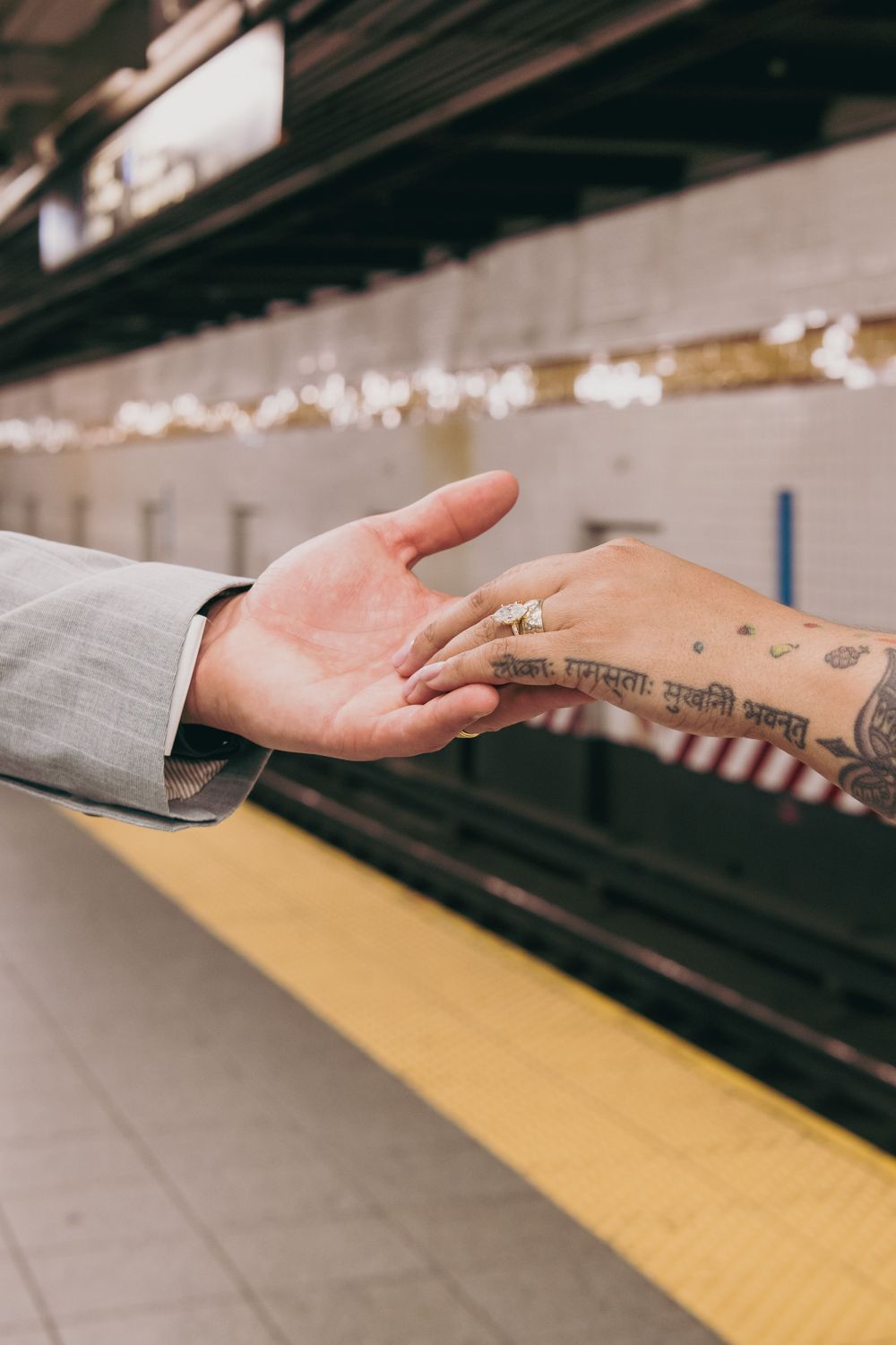 Couple holding hands at subway platform, brides tattooed hand wearing diamond engagement ring.