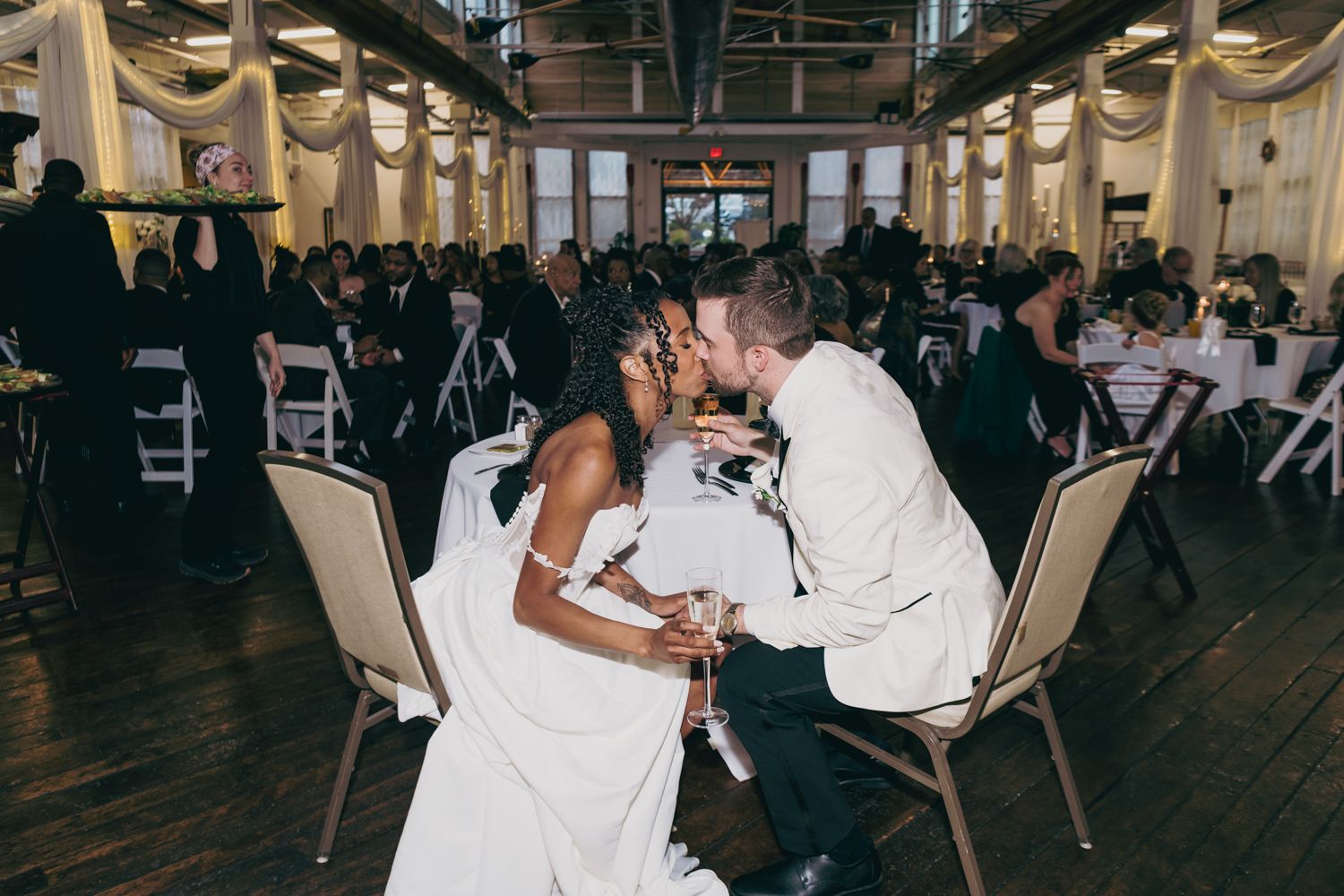 Newlyweds share a romantic kiss at their wedding reception in a warmly lit industrial venue.