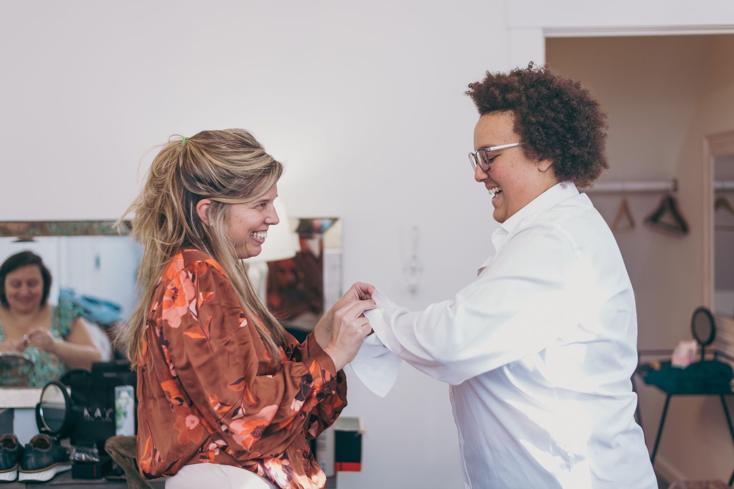 Two women smiling and sharing a joyful moment together during wedding preparations indoors.