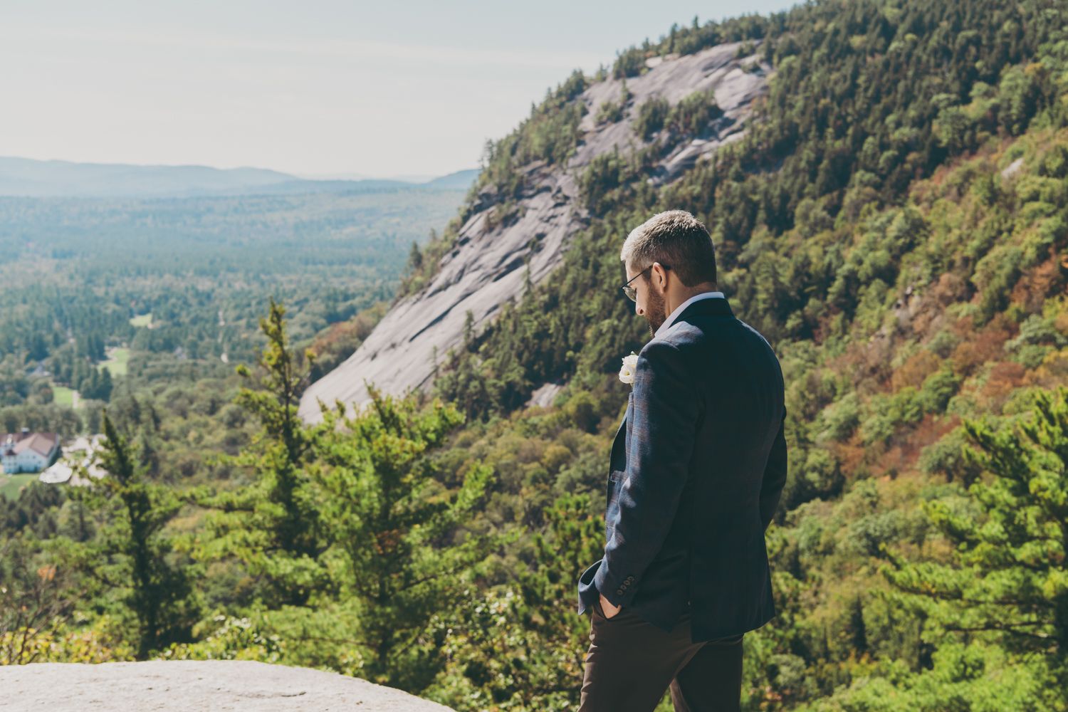 Groom in navy suit gazing at stunning mountain and forest landscape on his wedding day.