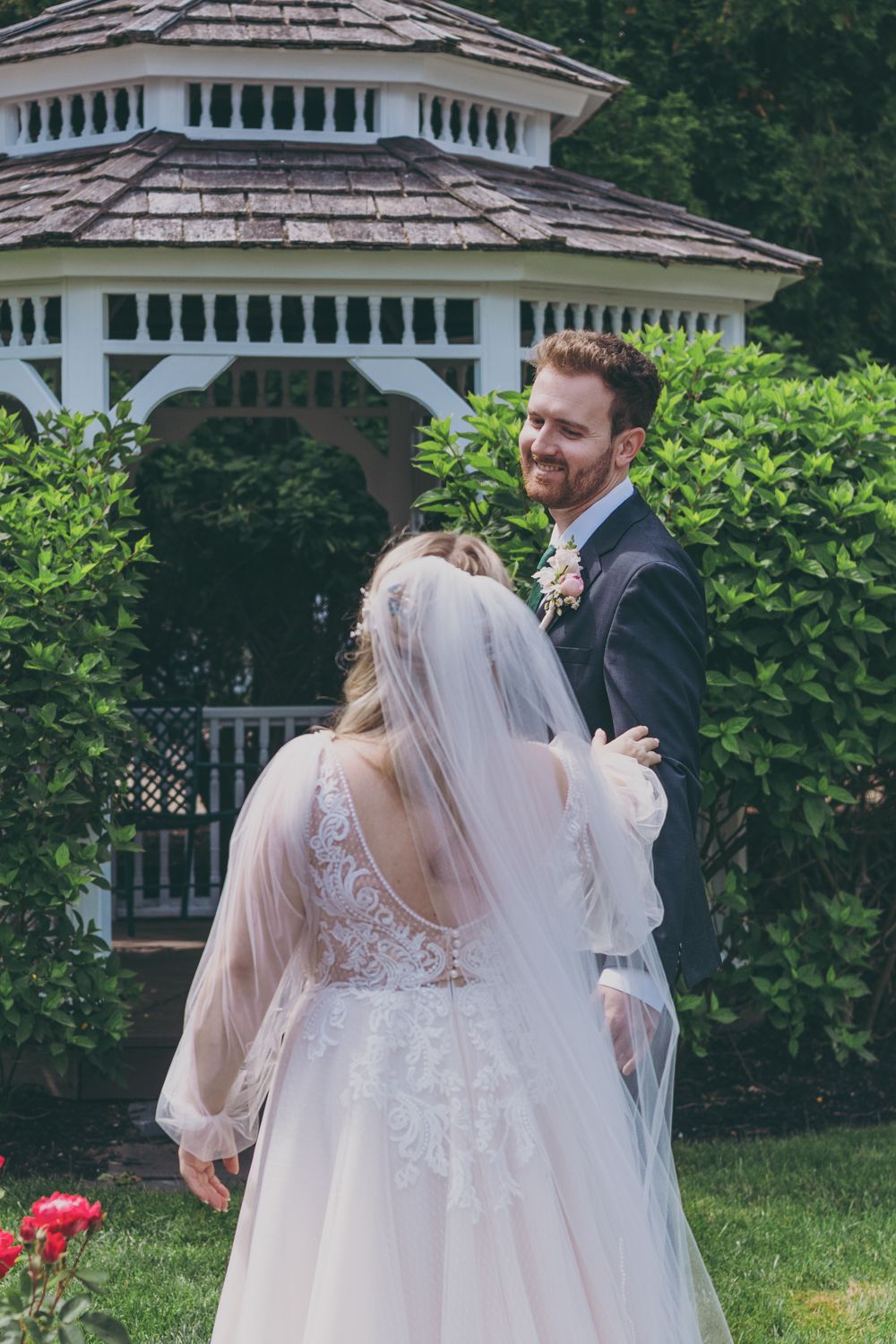 Groom in grey suit smiling at bride in lace wedding gown near white gazebo during first look.