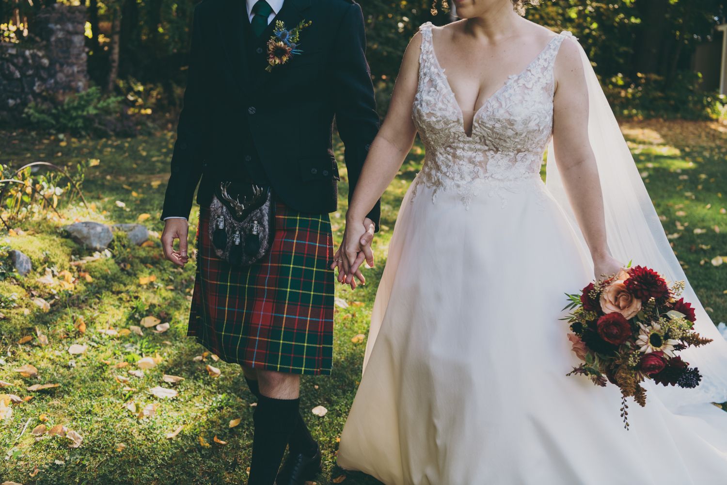 Bride and groom walking hand in hand, groom wearing traditional Scottish tartan kilt, outdoor wedding.