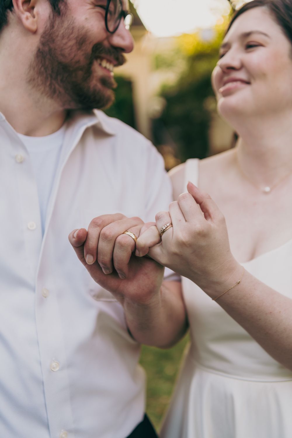 Newlyweds smile and hold hands showing off their wedding rings in a close-up outdoor portrait.