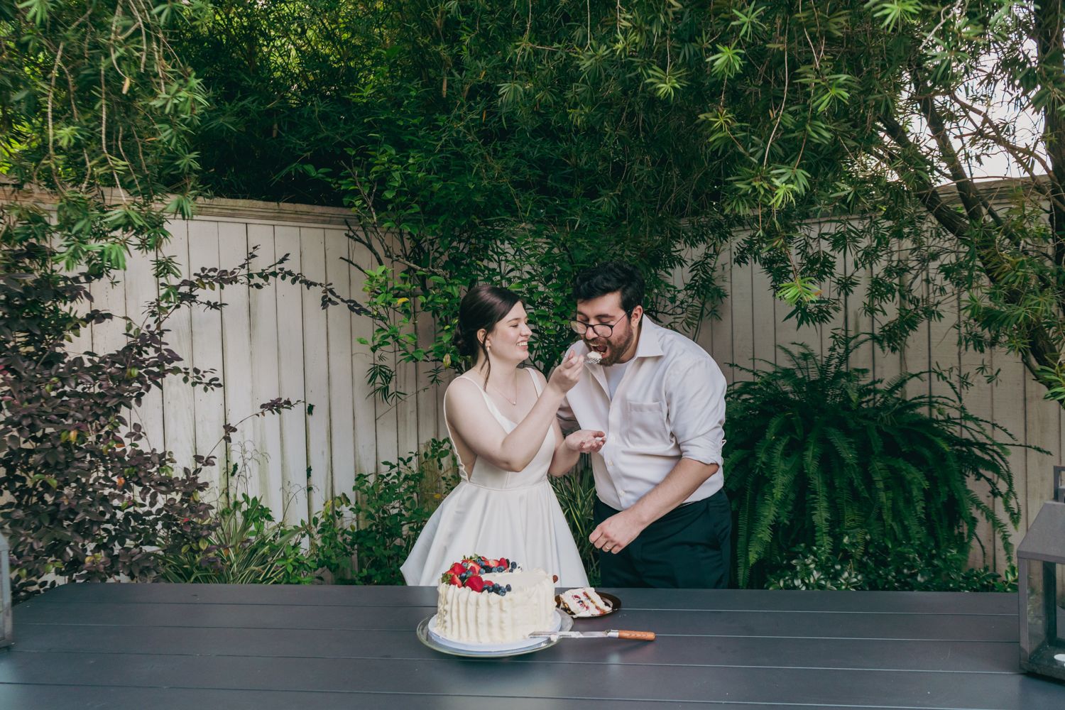 Bride feeds groom a bite of their white wedding cake decorated with fresh berries at outdoor reception.