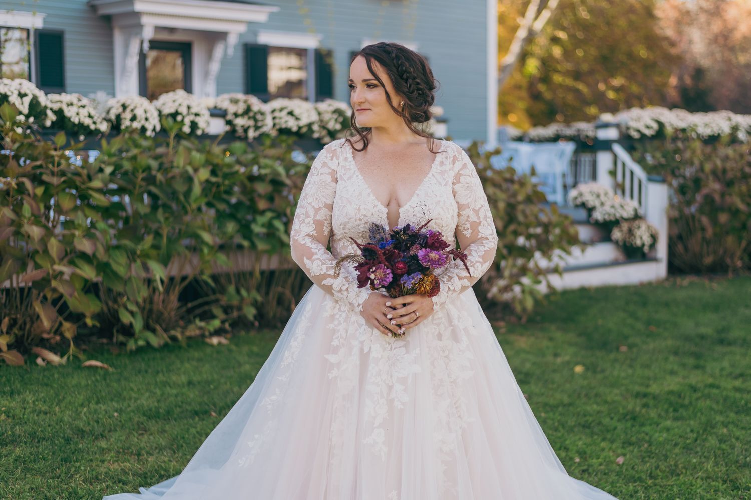 Bride in long-sleeve lace ball gown holds purple bouquet in front of white flower garden.