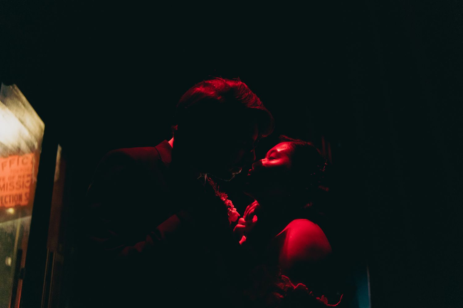 A couple shares a romantic kiss bathed in red neon light in a dark atmospheric setting.