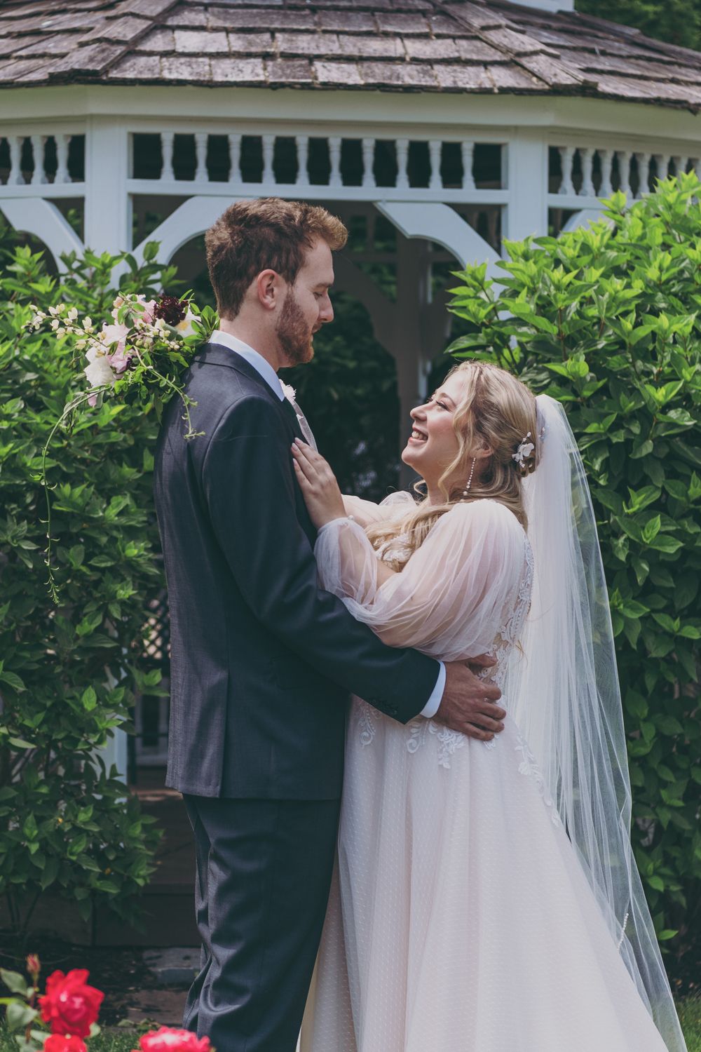 Laughing bride and groom embracing in front of white gazebo surrounded by green foliage.