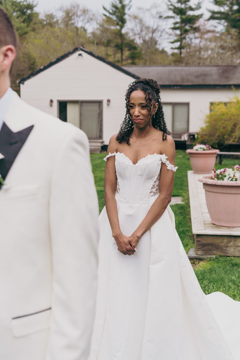 Bride in off-shoulder lace wedding gown gazes at groom during outdoor first look ceremony.