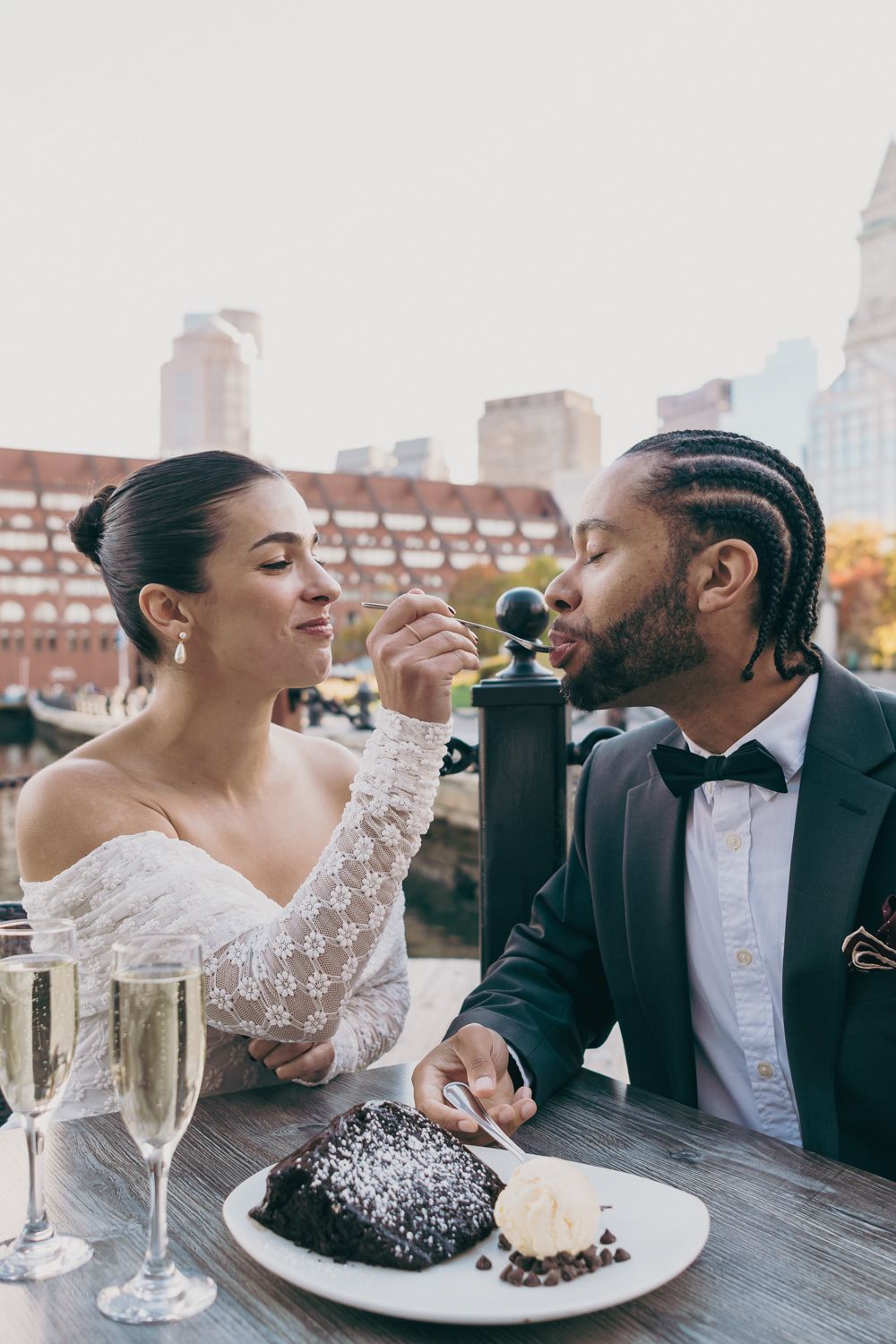 Newlywed couple sharing chocolate dessert and champagne at outdoor restaurant table.