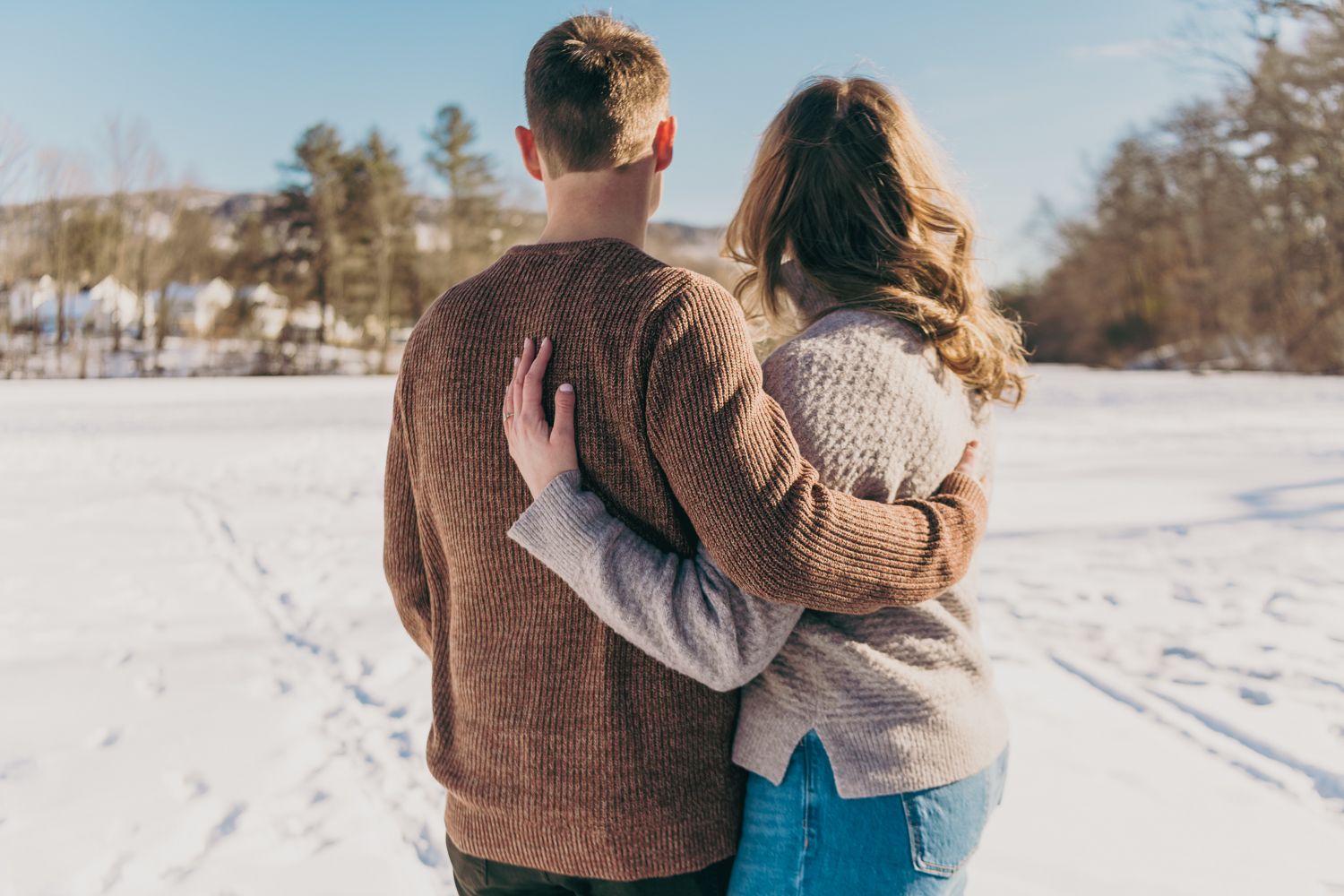 Couple embracing from behind in a snowy winter field surrounded by trees on a sunny day.