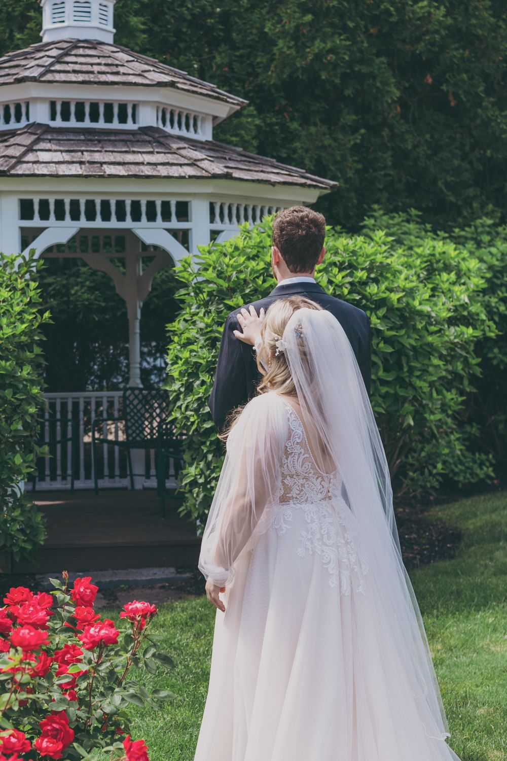 Bride and groom walking toward white garden gazebo surrounded by lush greenery and red roses.