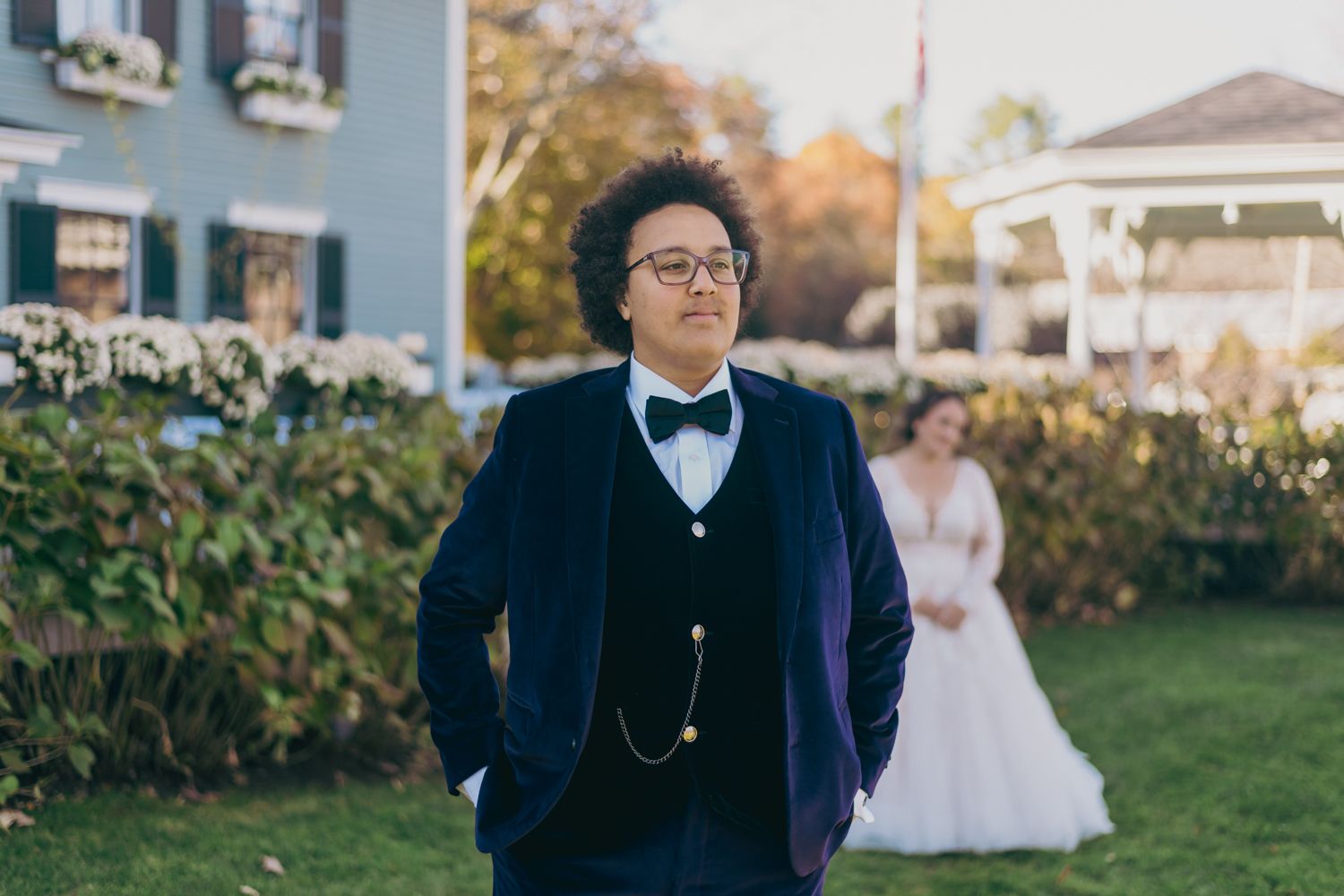 Groom in navy suit with bow tie and pocket watch stands outdoors before first look moment.