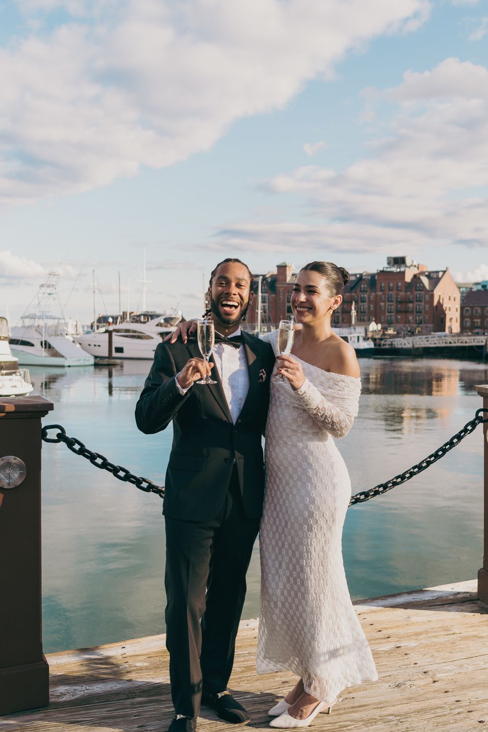 Happy interracial newlyweds toasting champagne on waterfront dock with marina in background.
