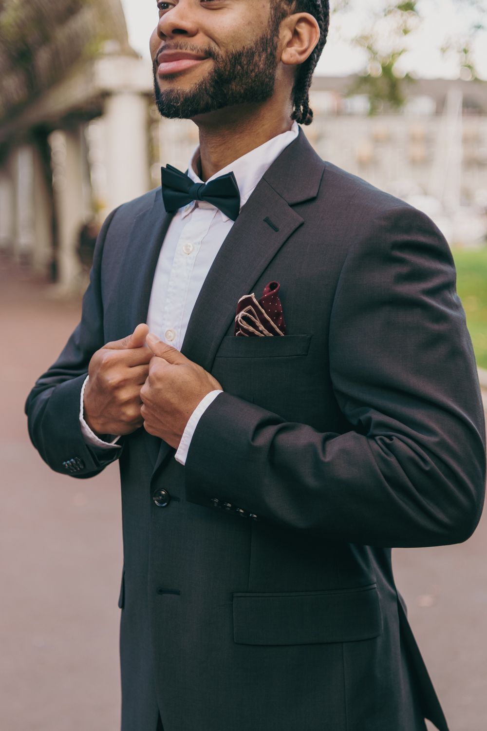 Elegant groom in black tuxedo with bow tie and burgundy paisley pocket square adjusting his jacket outdoors.