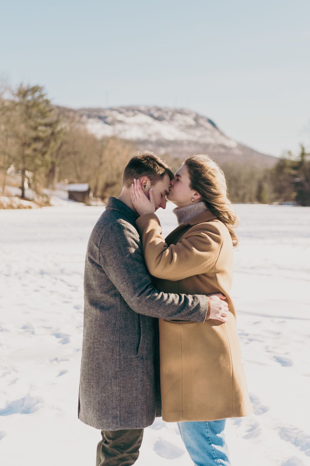 Couple sharing a romantic almost-kiss in a snowy winter landscape with mountains in the background.