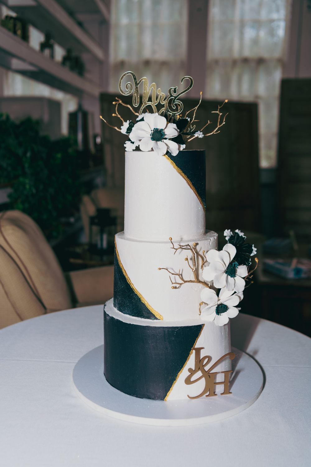 Elegant three-tier wedding cake with black and white fondant, gold accents, white flowers, and Mrs. cake topper.