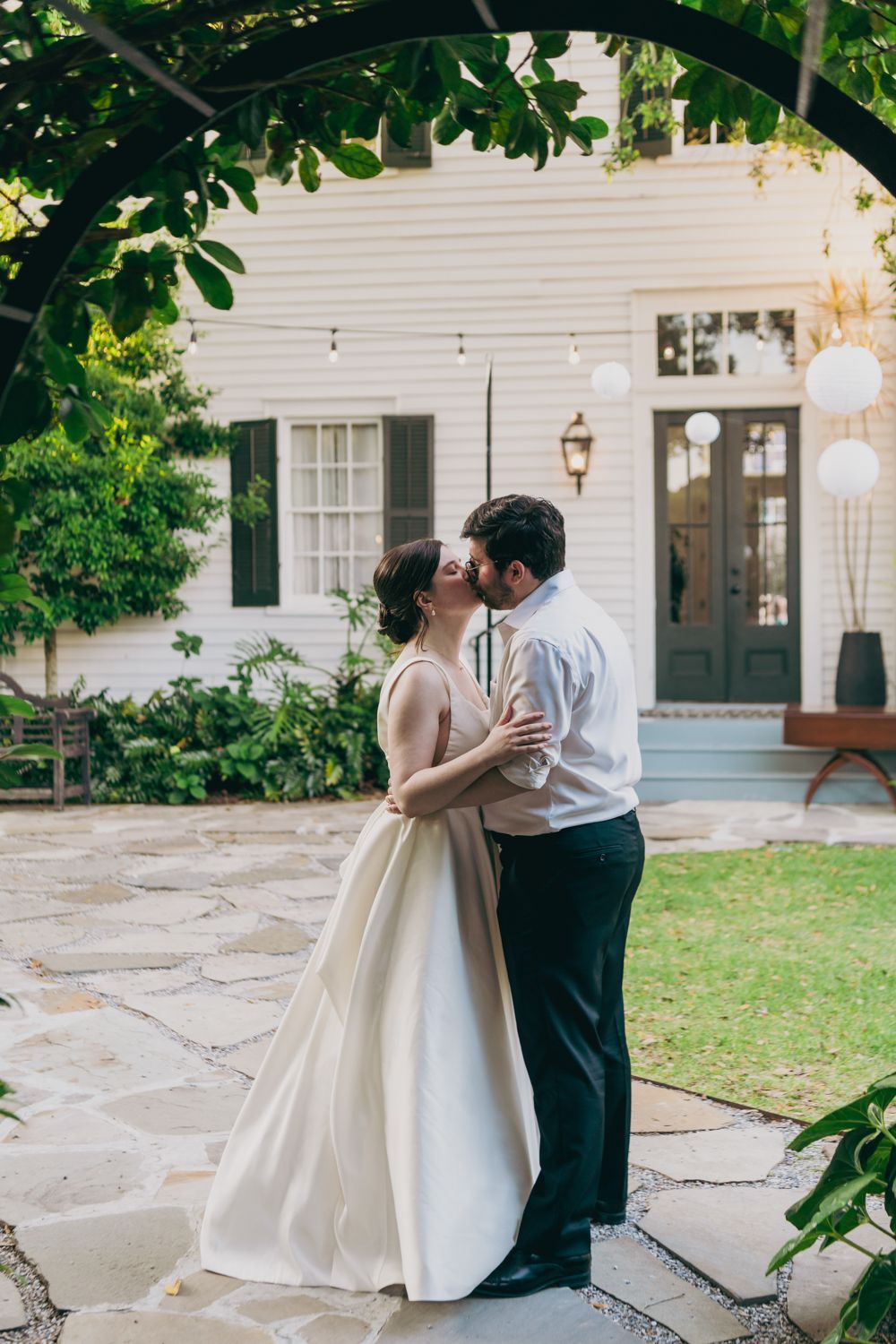 Bride and groom share a romantic kiss on a stone patio in front of a charming white colonial home.