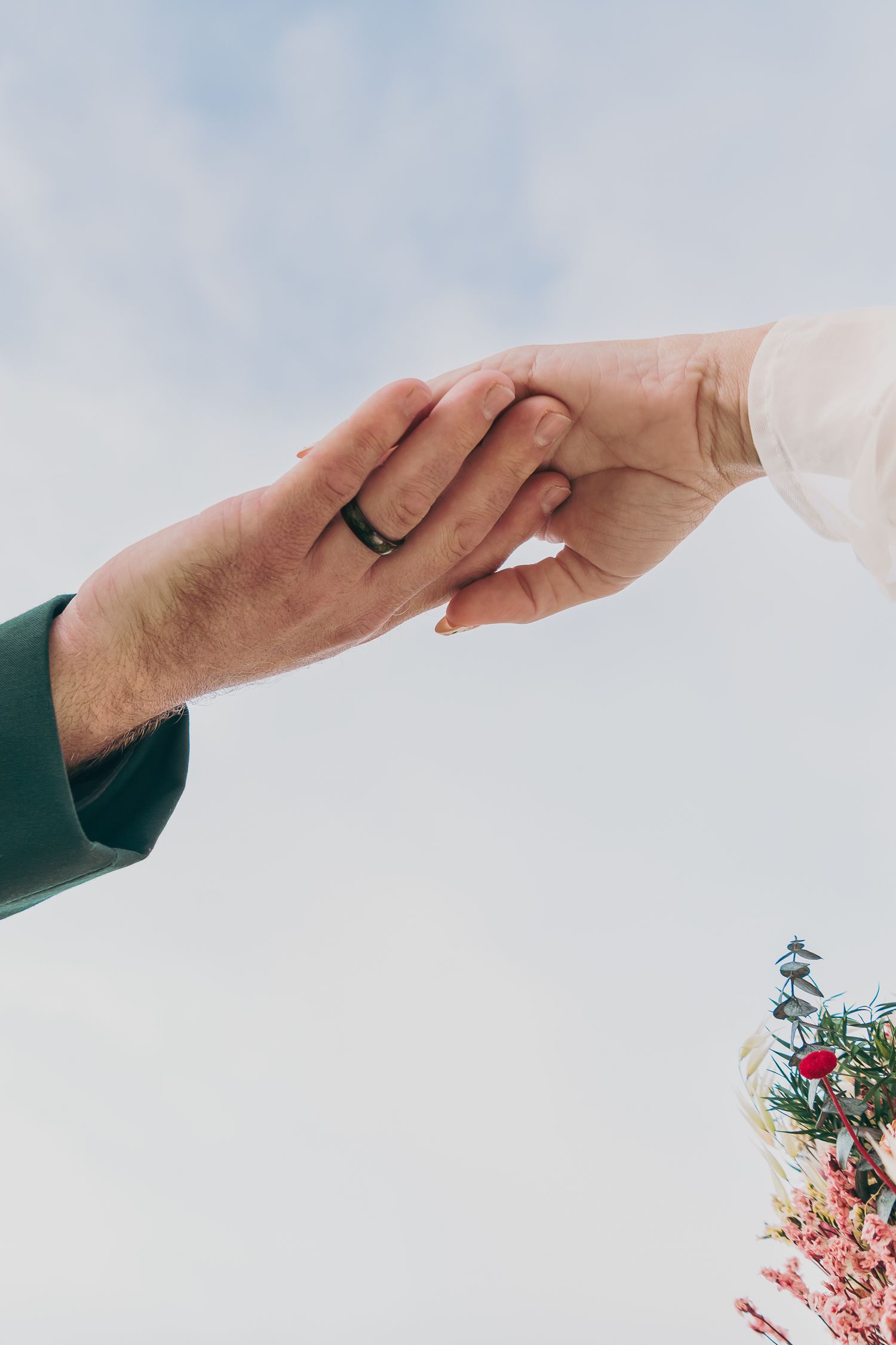 Two hands touching tenderly, one wearing a black wedding ring, with soft sky and flowers in background.