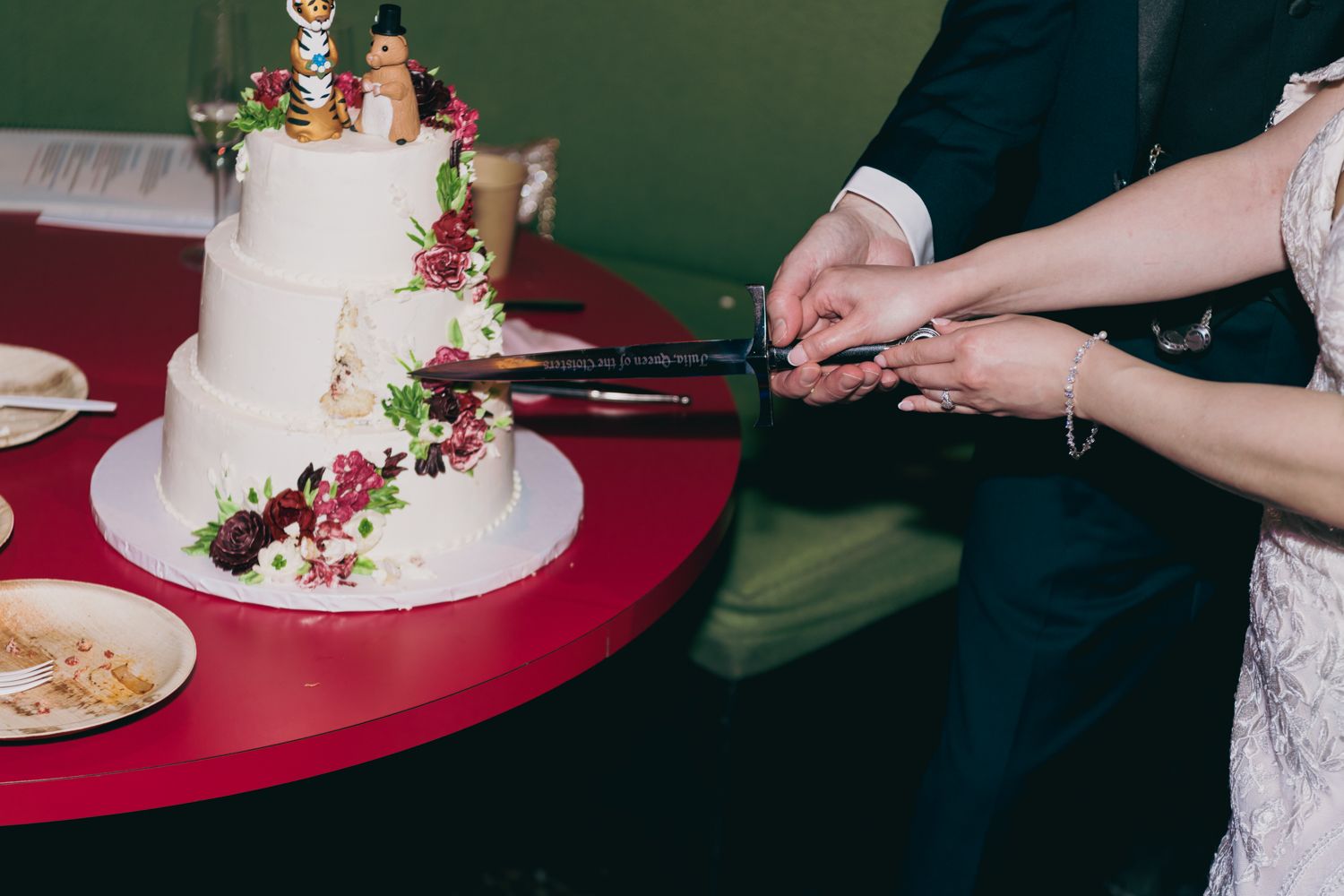 Bride and groom cutting their three-tier white wedding cake decorated with burgundy flowers on a red table.