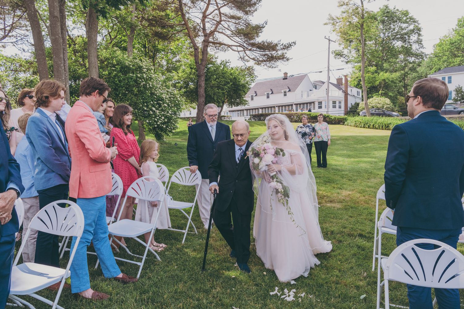 Bride walks down outdoor aisle escorted by elderly man with cane past seated wedding guests on lawn.