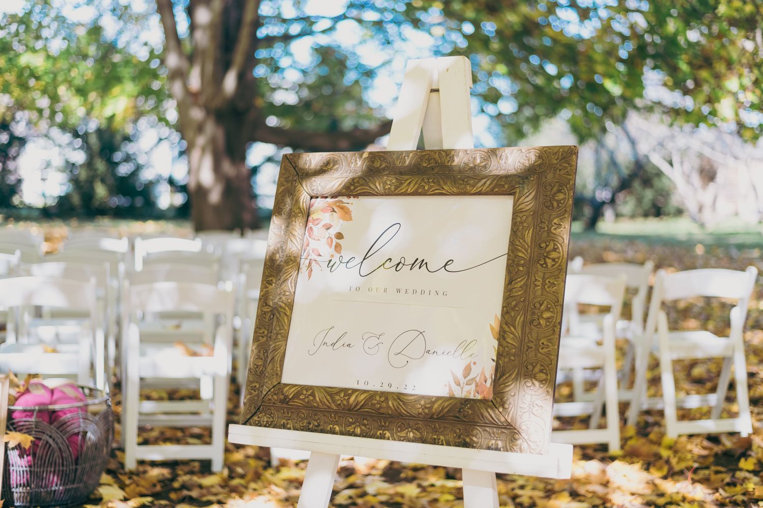 Elegant gold framed wedding welcome sign on easel at outdoor ceremony with white chairs and trees.