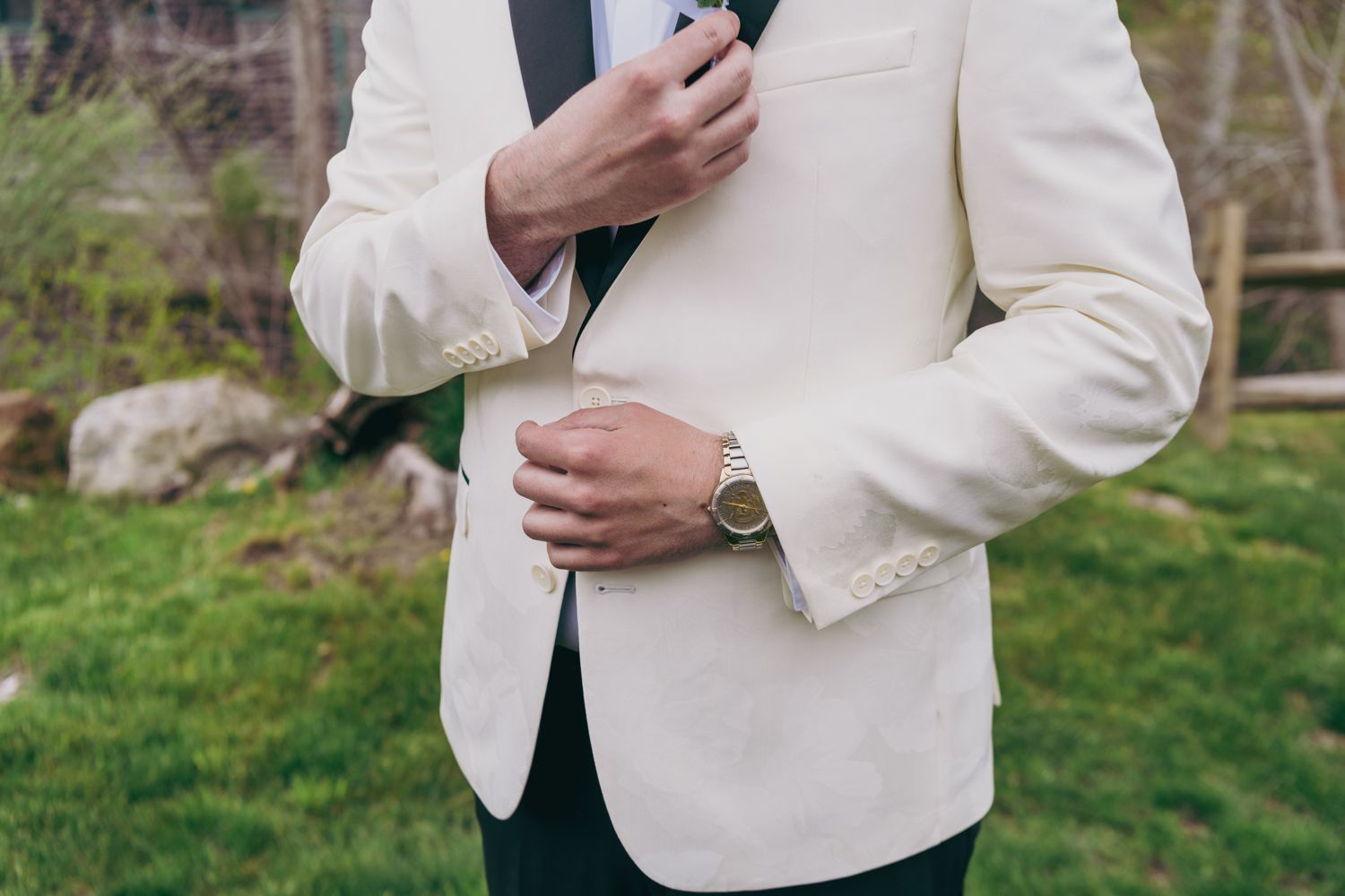 Groom in ivory white wedding blazer adjusting tie outdoors on wedding day.