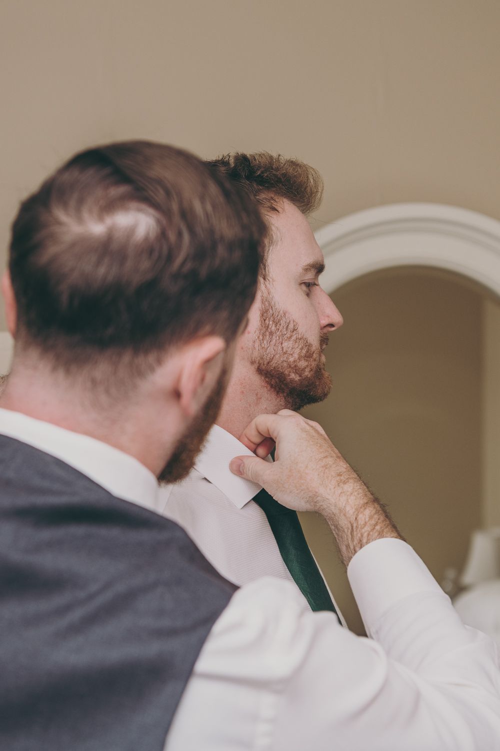 A groomsman helps the groom adjust his green tie while preparing for a wedding ceremony.