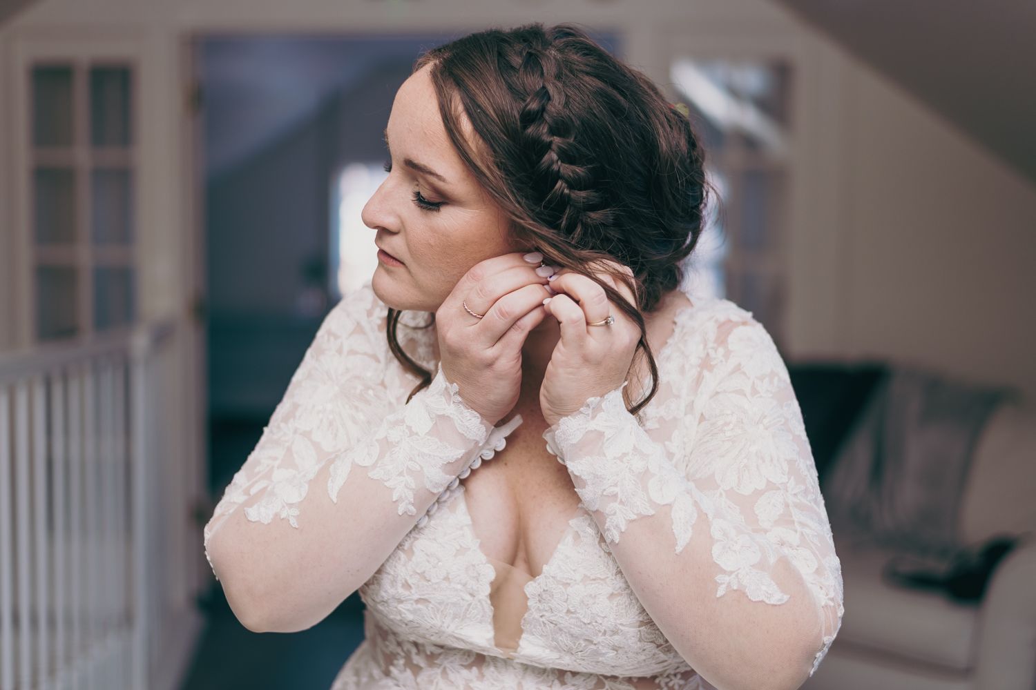 Bride in lace wedding dress putting on earrings with braided updo hairstyle.