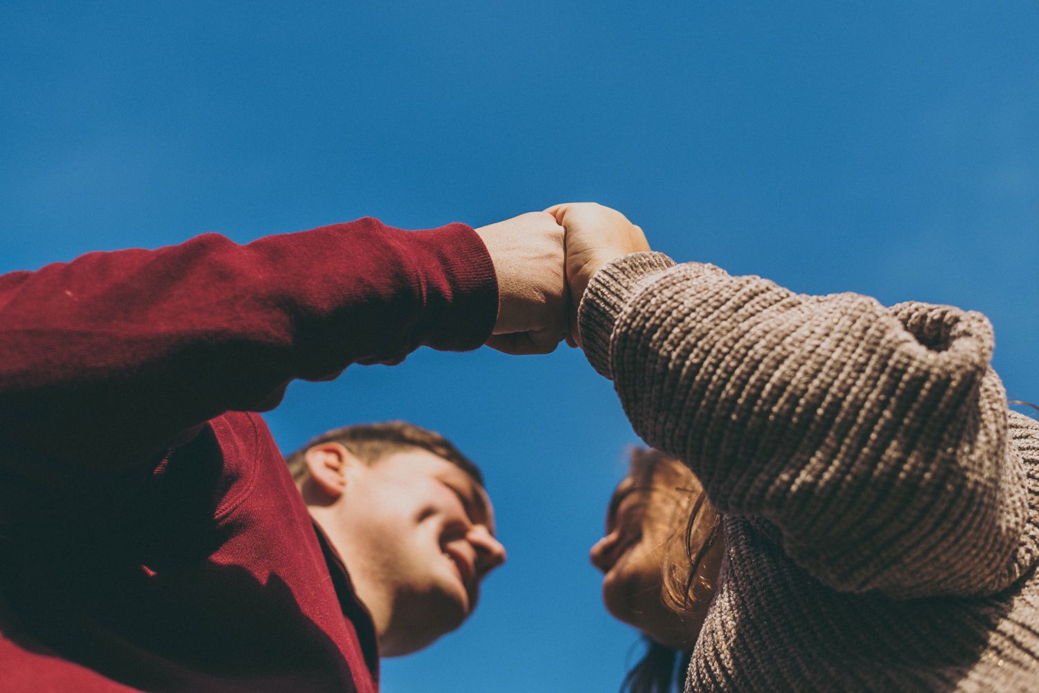Two people fist bumping against a clear blue sky, one in a red hoodie and the other in a beige knit sweater.