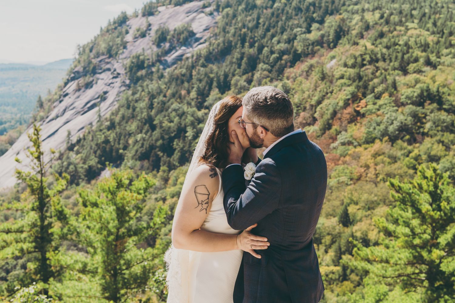 Newlywed couple shares first kiss with lush mountain forest backdrop.