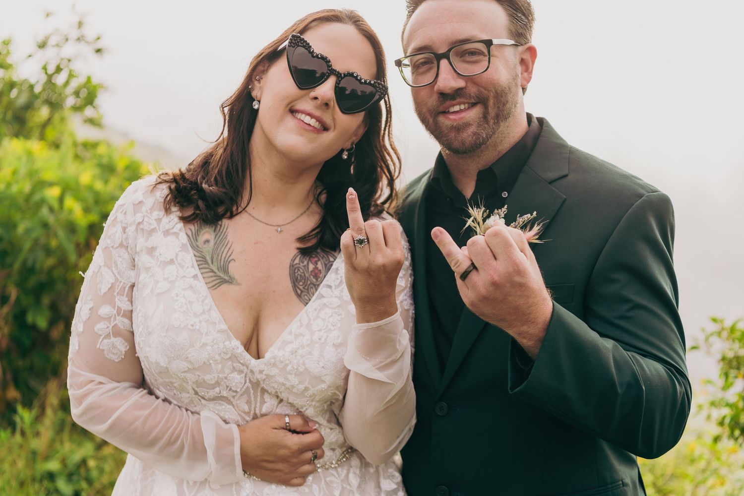 Bride and groom smiling and showing off wedding rings outdoors on a misty hillside, bride in lace dress with sunglasses.