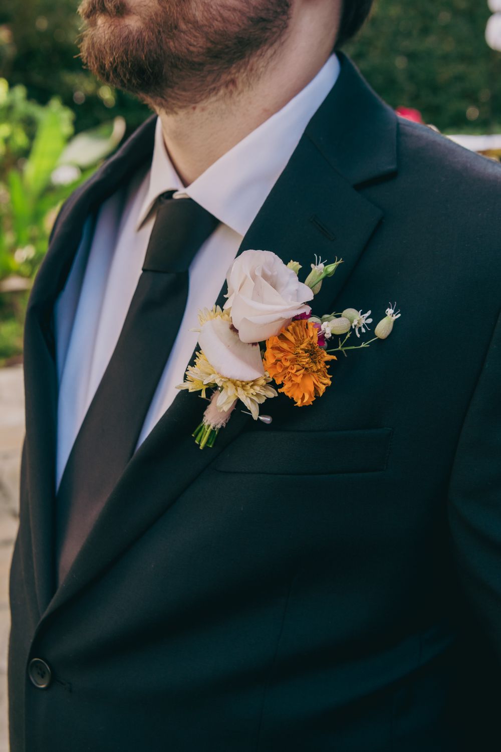 Close-up of groom's boutonniere featuring a blush rose, orange zinnia, and delicate white flowers on dark suit.