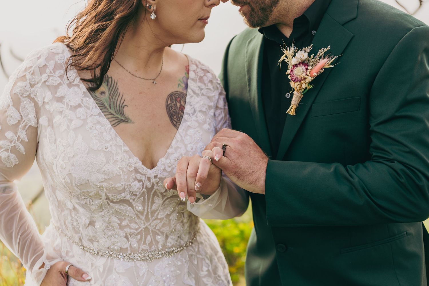 Bride and groom exchanging rings, bride in lace gown with tattoo, groom in green suit with floral boutonniere.