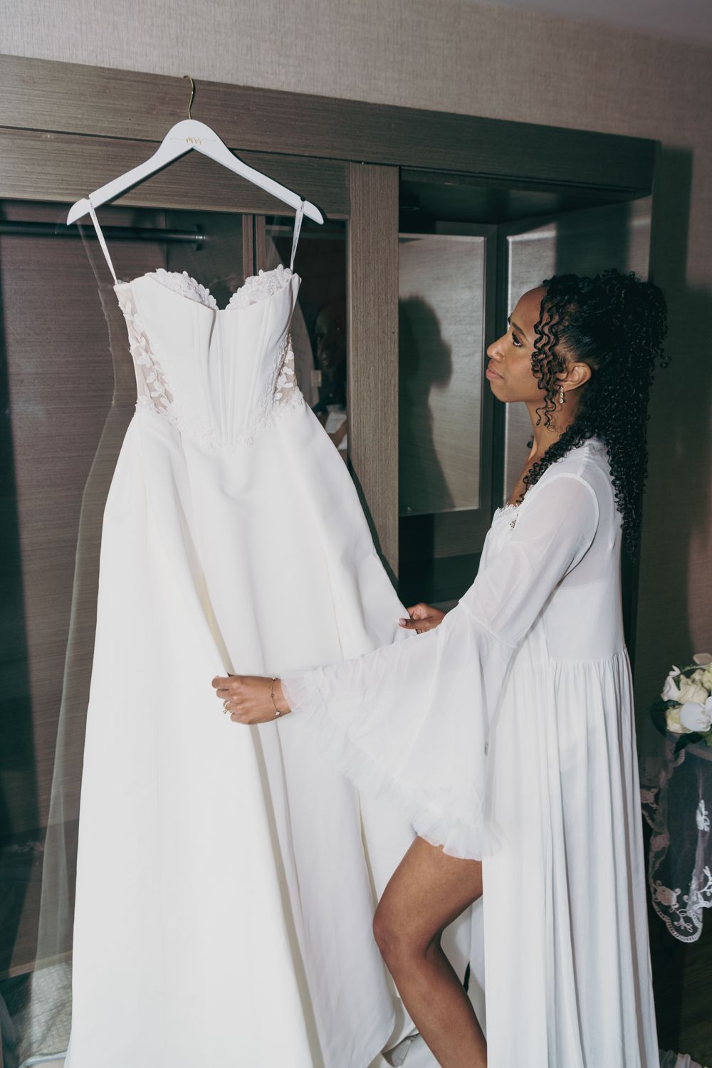 Bride in white robe admiring her wedding dress on a hanger in a hotel room before the ceremony.