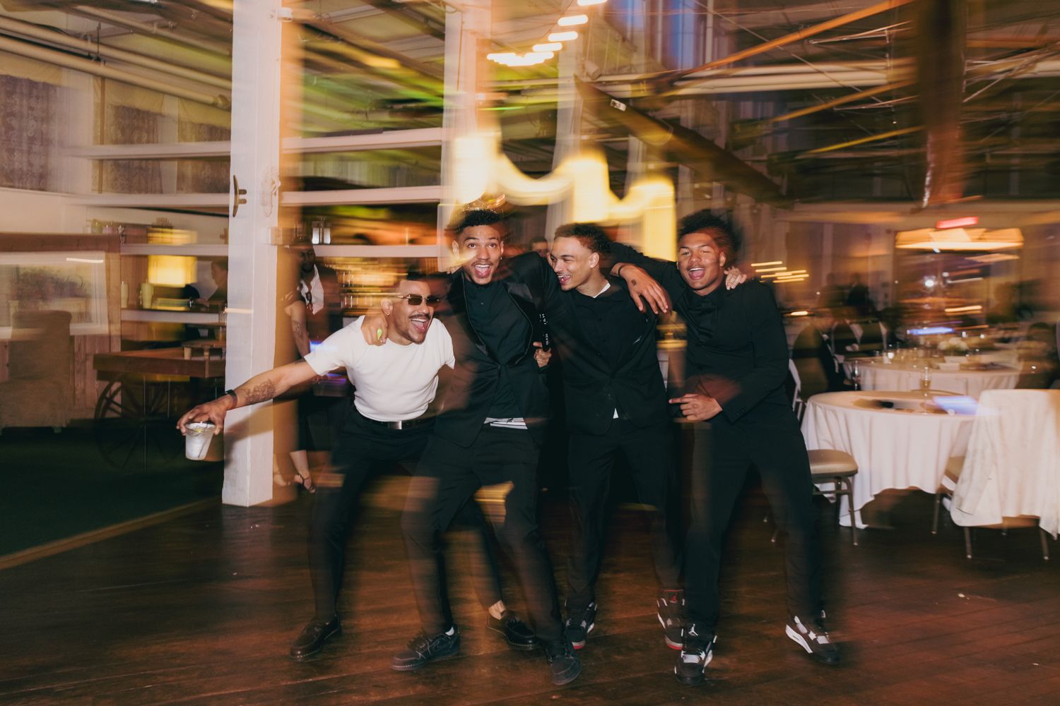 Four men celebrating and dancing together on wedding reception dance floor with bokeh lighting effect.