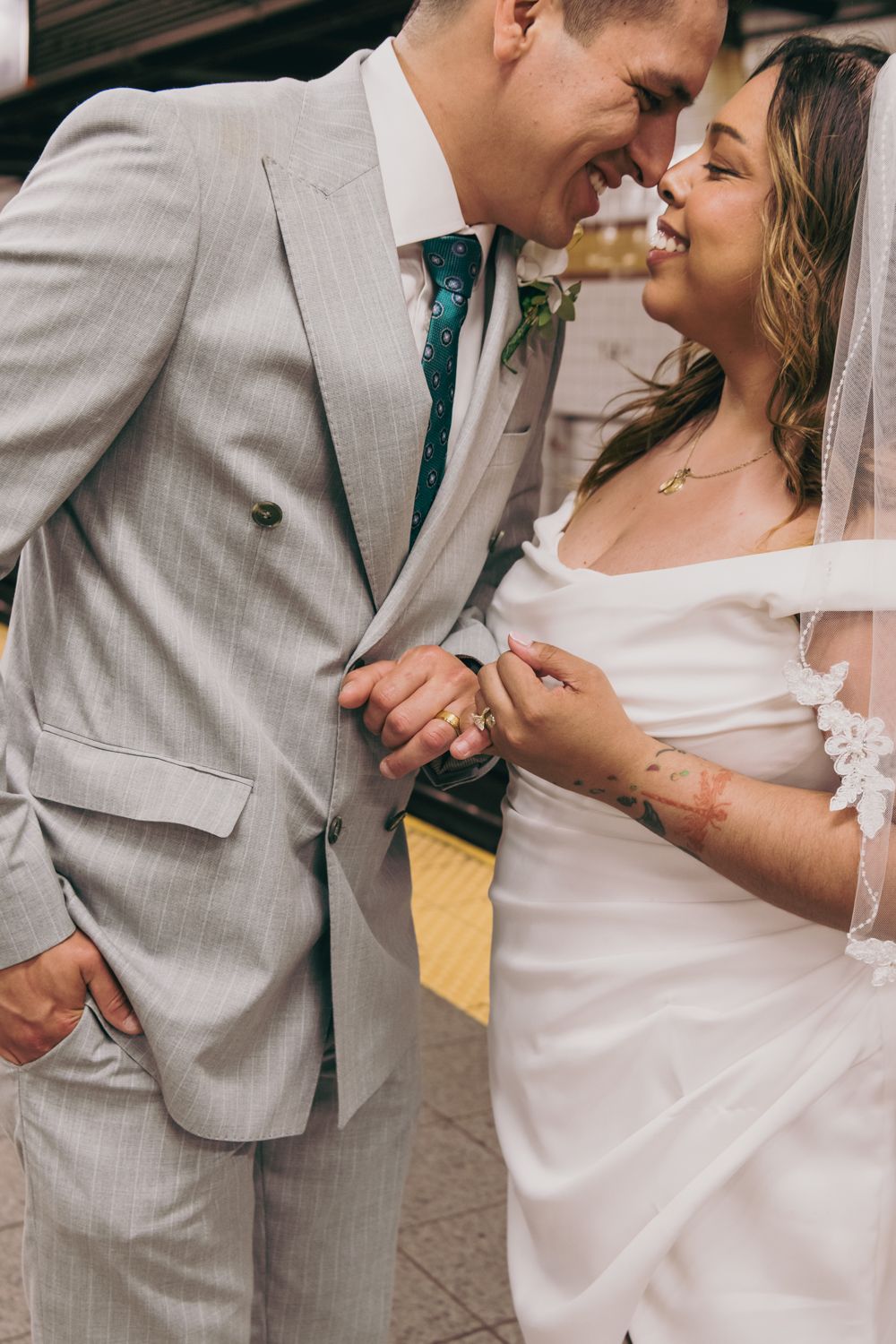 Bride in white gown with lace veil and tattooed groom in gray suit holding hands and smiling at each other.