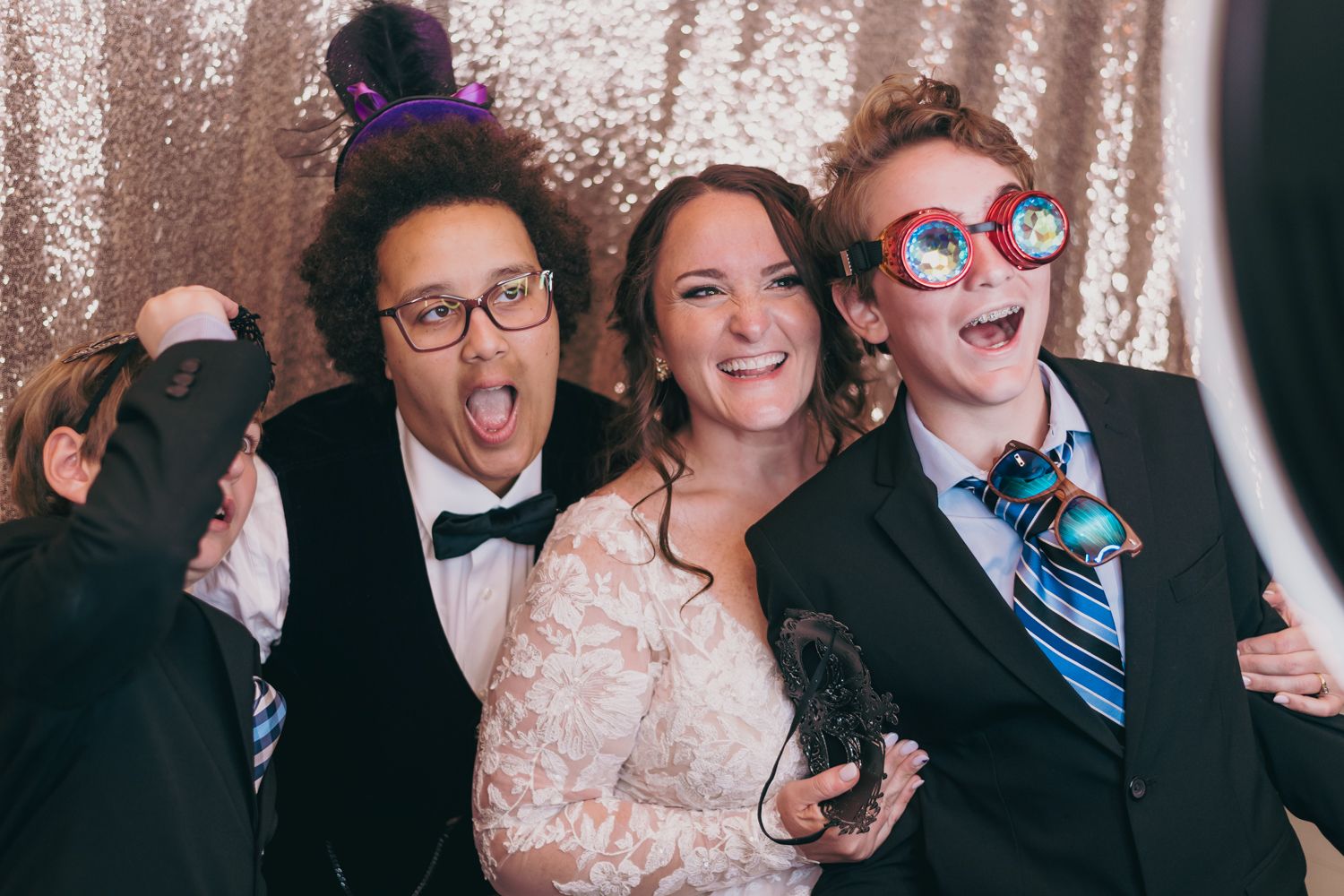 Three people posing fun at a wedding photo booth with sparkly backdrop, wearing silly glasses and props.