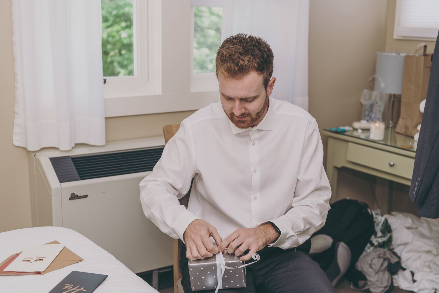 Groom in white shirt opening a gift box while seated at a table in a bright room before his wedding.