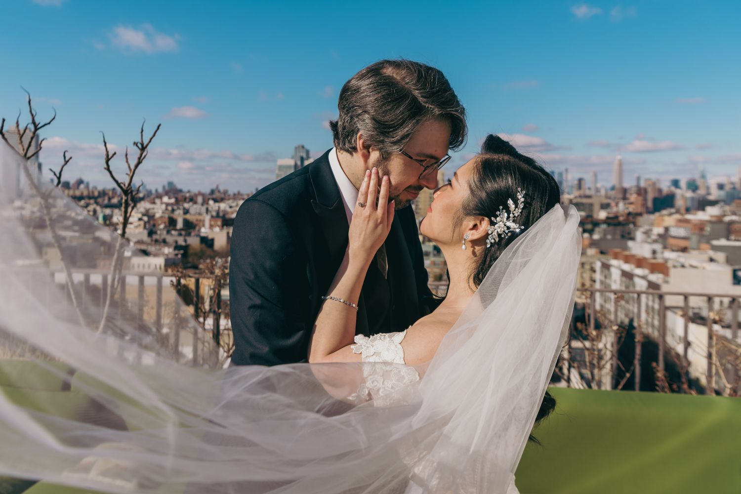 A bride and groom share a romantic kiss on a rooftop with a city skyline backdrop, her veil flowing in the wind.