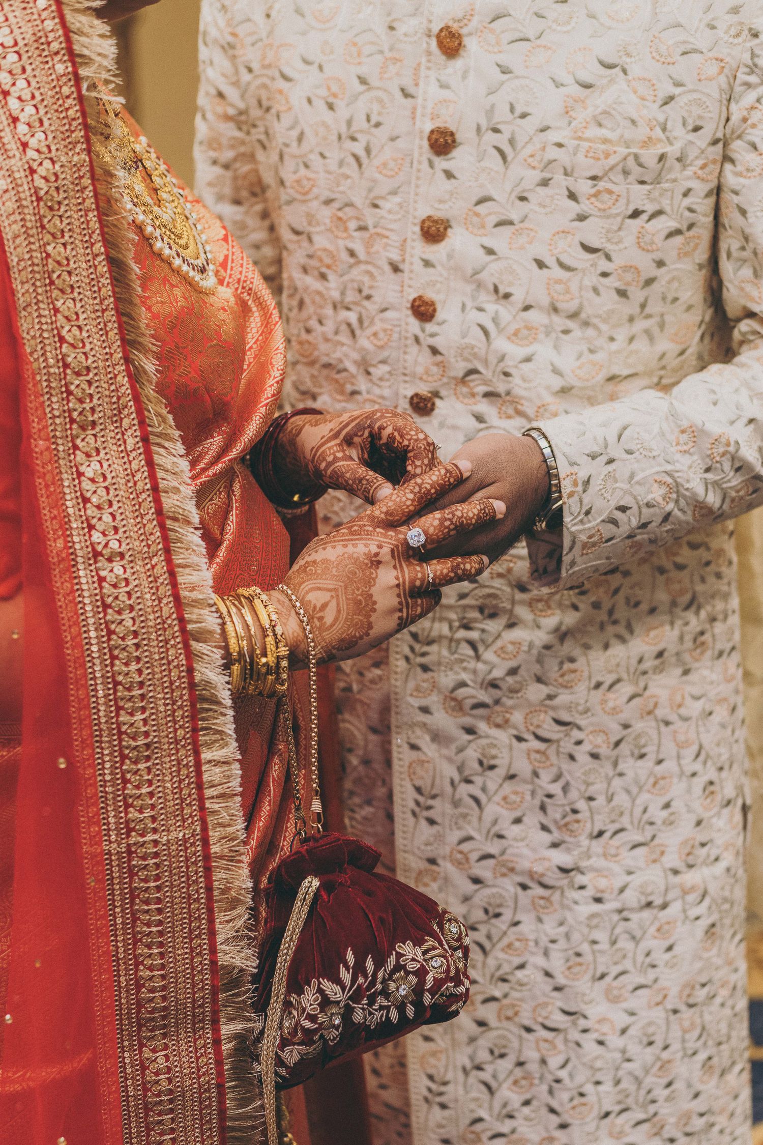 Indian bride and groom exchanging rings, bride wearing mehndi, gold bangles, red saree, groom in white embroidered sherwani.